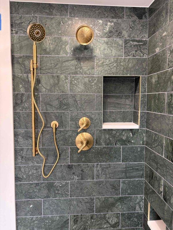 Completed installation of brushed gold shower fixture set in remodeled shower enclosure with dark grey marble-effect tile. System includes wall-mounted fixed shower head, handheld shower head with slide bar, dual valve handles, and central mixing valve/diverter, all in matching brushed gold finish. Payment collected upon completion of sale.