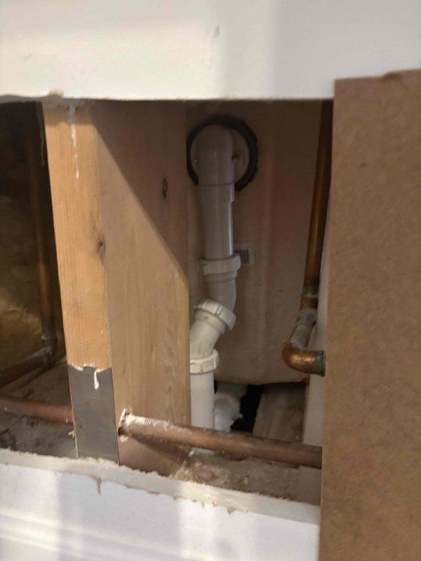 Responded to call for active water leak beneath bathtub in master bedroom. Accessed plumbing through access panel and located leak source on white PVC drain pipe running vertically in wall cavity between master bathtub and adjacent closet. Documented plumbing configuration showing P-trap assembly with black rubber coupling and standard white PVC drainage components. Photographed affected area revealing drain pipe connections, copper supply lines, and cabinet interior to verify leak location and assess extent of water damage for necessary repairs.