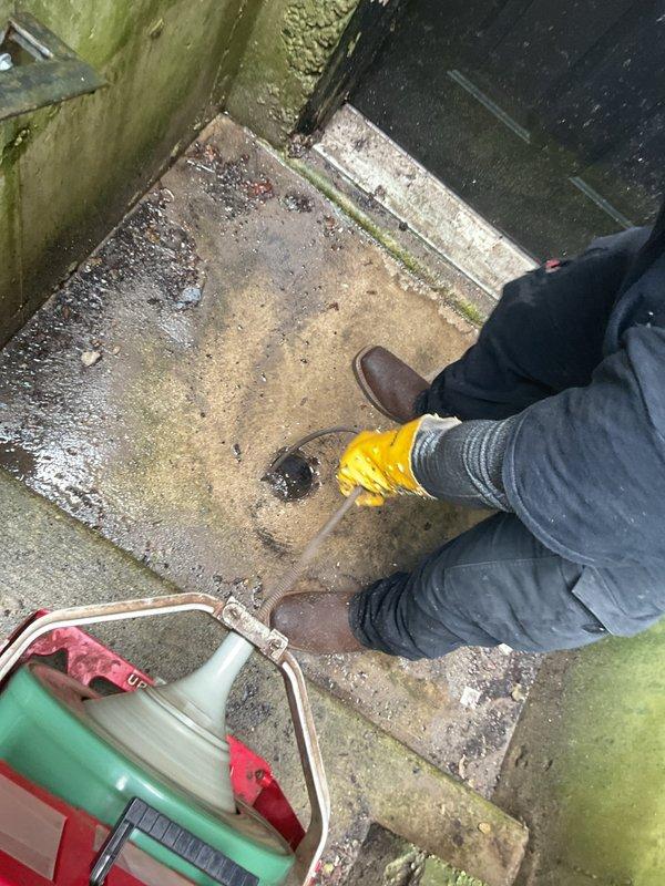 Utilized main-line cable machine from ground-level cleanout to rod and clear main sewer line, performing courtesy camera inspection to verify proper flow. Successfully rodded floor drain at rear of building to restore drainage functionality. Work completed in basement area with standing water conditions present, requiring navigation of damp environment to access drainage points.