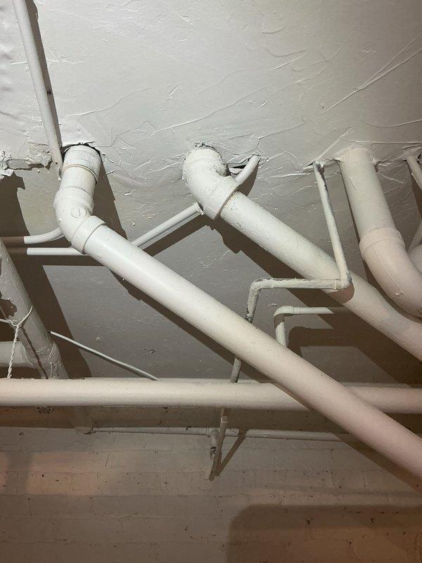 Completed kitchen stack repair involving extensive PVC drainage piping system in basement utility area. Repaired and reconfigured multiple sections of white PVC drain lines including horizontal ceiling runs and vertical drops, with proper mounting brackets and support hangers installed throughout. Work included addressing junctions, elbows, and connections serving the double-basin utility sink and associated plumbing fixtures to restore proper drainage function.