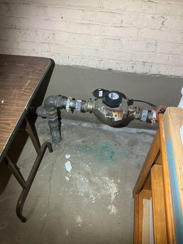 Responded to service request for sewer camera inspection due to sink hole in front of building. Performed comprehensive video camera inspection of main sewer line from house connection to street to assess condition and identify potential causes of ground subsidence. Documentation photos captured water meter assembly location and foundation area showing structural concerns with visible concrete deterioration, cracking, and debris accumulation beneath building structure.