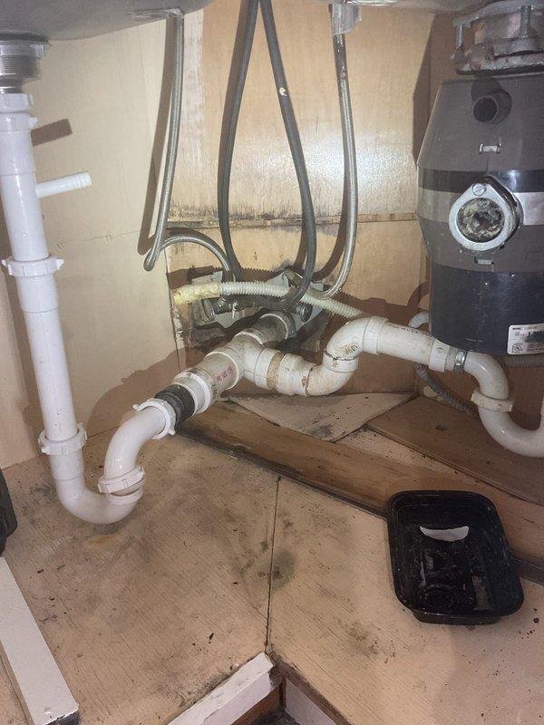 Responded to a kitchen sink drain assembly leak beneath a double-basin sink in a 120-year-old home. Upon inspection, identified leaking from PVC drain piping connected to the P-trap assembly and garbage disposal unit, with visible water staining on cabinet interior surfaces. Documented existing plumbing configuration including white PVC drainage system, garbage disposal connections, and flexible supply lines, then performed necessary repairs to eliminate the leak and restore proper drainage function.
