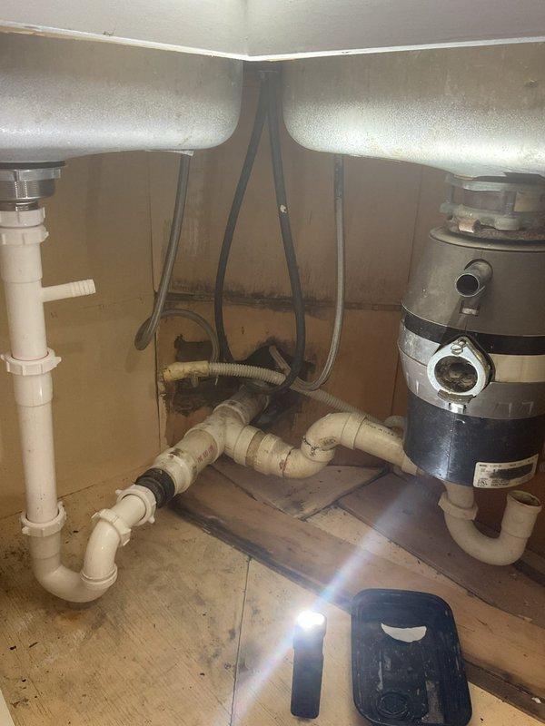 Responded to a kitchen sink drain assembly leak beneath a double-basin sink in a 120-year-old home. Upon inspection, identified leaking from PVC drain piping connected to the P-trap assembly and garbage disposal unit, with visible water staining on cabinet interior surfaces. Documented existing plumbing configuration including white PVC drainage system, garbage disposal connections, and flexible supply lines, then performed necessary repairs to eliminate the leak and restore proper drainage function.