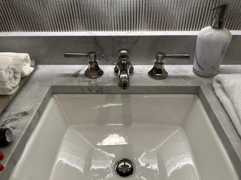 Installed three-piece chrome faucet assembly with lever-style handles and center spout on gray marble countertop with undermount white basin. Connected braided stainless steel supply lines to existing copper supply pipes and installed pop-up drain assembly with stopper mechanism. Return visit required to address improper installation of faucet handles and drain pull mechanism that did not meet quality standards.