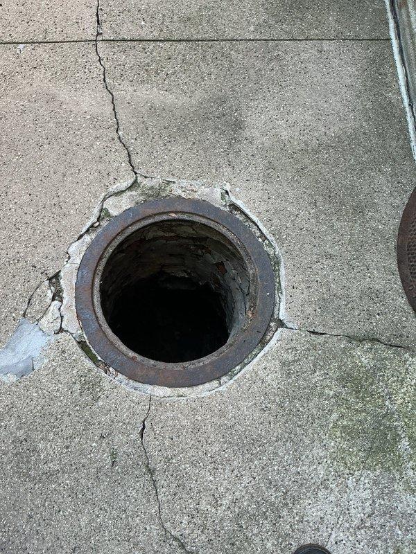 Conducted site assessment for catch basin rebuild in residential courtyard area. Inspection revealed deteriorated circular drain opening with significant concrete cracking radiating from the fixture and along surrounding surface. Metal drain collar exhibited extensive rust and weathering with brownish-red corrosion around the rim. Concrete showed multiple stress fractures, surface deterioration, and chipping at drain interface points. White sealant material visible along drain perimeter indicated previous repair attempts. Documented existing drainage infrastructure including four lower-level access points protected by metal railing, circular drain cover in center courtyard, and overall paving slab condition. Photographed evidence of water management issues, structural concrete degradation, and compromised drain integrity for estimate preparation. Evaluated scope of work required for complete catch basin reconstruction including removal of existing deteriorated fixture, concrete repair,