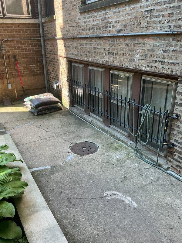 Conducted site assessment for catch basin rebuild in residential courtyard area. Inspection revealed deteriorated circular drain opening with significant concrete cracking radiating from the fixture and along surrounding surface. Metal drain collar exhibited extensive rust and weathering with brownish-red corrosion around the rim. Concrete showed multiple stress fractures, surface deterioration, and chipping at drain interface points. White sealant material visible along drain perimeter indicated previous repair attempts. Documented existing drainage infrastructure including four lower-level access points protected by metal railing, circular drain cover in center courtyard, and overall paving slab condition. Photographed evidence of water management issues, structural concrete degradation, and compromised drain integrity for estimate preparation. Evaluated scope of work required for complete catch basin reconstruction including removal of existing deteriorated fixture, concrete repair,