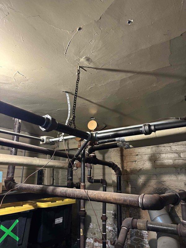 Accessed concealed plumbing system through existing wall opening to address repair requirements. Removed section of drywall to expose interior wall cavity and gain access to vertical piping runs between wall studs. Inspected metallic conduit and plumbing infrastructure within the wall assembly.

Evaluated extensive basement ceiling piping network including multiple horizontal supply and drain lines of various diameters. Assessed condition of metallic and aged pipe sections, support hangers, and mounting straps. Inspected pipe joints, connections, and shut-off valve access points throughout the mechanical space. Documented existing infrastructure condition including areas of surface deterioration and corrosion on older pipe sections for repair recommendations.