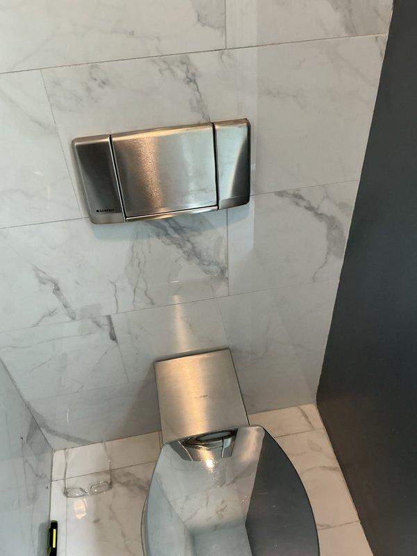 Responded to service call for two toilet issues. Inspected first toilet with reported leak below and assessed second toilet with water leaking at base onto floor. Evaluated wall-mounted flush system and contemporary toilet fixture in marble-tiled bathroom. Documented conditions of both units and surrounding areas, including examination of potential seal failures and base leakage points requiring repair or replacement.