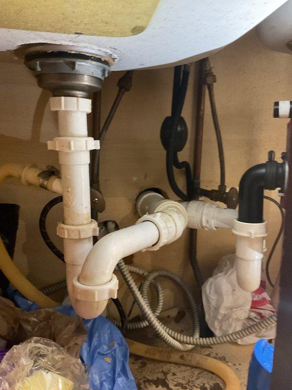 Responded to service call for double-basin kitchen sink with standing water backup and overflow issues. Upon inspection, found both basins retaining water with visible debris, indicating drainage obstruction in the common waste line or P-trap assembly. Accessed under-sink plumbing configuration which included white PVC P-trap, garbage disposal unit with associated drain lines, and dishwasher connection hose. Disassembled P-trap compression fittings to inspect for blockage accumulation. Cleared obstruction from trap assembly and drain line, removing accumulated food particles, grease buildup, and debris causing the backup. Verified proper water flow through both basin drains and disposal unit. Reassembled all plumbing connections, ensuring proper sealing at compression fittings to prevent future leaks. Tested system under full water flow to confirm complete drainage restoration and verified no leaks at connection points. System returned to normal operation with both basins draining free