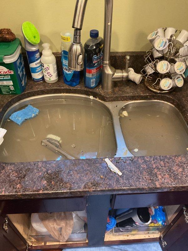 Responded to service call for double-basin kitchen sink with standing water backup and overflow issues. Upon inspection, found both basins retaining water with visible debris, indicating drainage obstruction in the common waste line or P-trap assembly. Accessed under-sink plumbing configuration which included white PVC P-trap, garbage disposal unit with associated drain lines, and dishwasher connection hose. Disassembled P-trap compression fittings to inspect for blockage accumulation. Cleared obstruction from trap assembly and drain line, removing accumulated food particles, grease buildup, and debris causing the backup. Verified proper water flow through both basin drains and disposal unit. Reassembled all plumbing connections, ensuring proper sealing at compression fittings to prevent future leaks. Tested system under full water flow to confirm complete drainage restoration and verified no leaks at connection points. System returned to normal operation with both basins draining free
