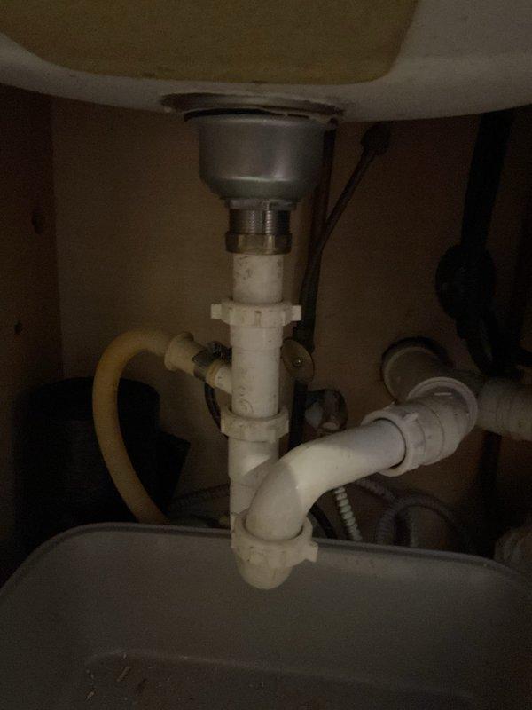 Inspected kitchen sink drain assembly and P-trap plumbing connections beneath basin. Documented existing drain basket strainer, white PVC P-trap assembly with slip-joint fittings, and associated drainage piping showing mineral deposits and age-related discoloration. Verified proper trap configuration for sewer gas prevention and assessed overall condition of under-sink plumbing components.
