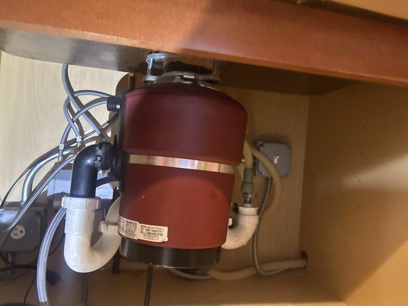 Responded to water leak between kitchen sink and garbage disposal. Upon inspection, located failed garbage disposal mounting flange assembly showing severe corrosion and structural deterioration. The mounting flange exhibited extensive pitting, material breakdown, and complete cracking of the gasket seal, which was the source of the active leak. Documented the damaged components and existing plumbing configuration under the sink, including disposal unit connections and drainage system. Advised on necessary repairs to eliminate the leak and prevent further water damage to the cabinet area.