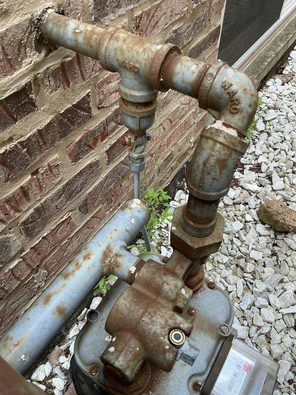Responded to gas leak reported near water heater and furnace area. Upon arrival, conducted comprehensive visual inspection of gas piping system in mechanical room and exterior gas meter assembly. Located active leak at corroded gas piping connections near water heater installation. Performed pressure test on gas system to verify leak location and assess system integrity. Documented heavily corroded exterior gas meter piping and deteriorated connections showing significant rust accumulation and compromised fittings. Identified multiple shut-off valves with operational concerns due to age and corrosion. Photographed interior gas piping configuration including black iron pipes, brass fittings, and safety valves for documentation purposes. Exterior gas supply assembly showed extensive weathering with compromised pipe coating and joint integrity requiring immediate attention to prevent further gas escape and ensure safe operation of gas-fired appliances.