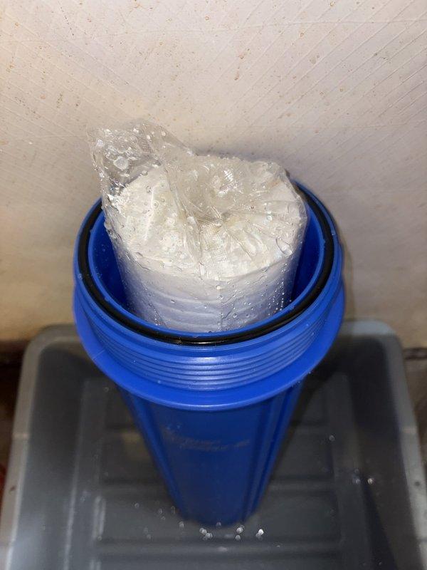 Inspected water filtration system and documented condition of blue filter housing with white pleated cartridge. Filter housing showed moisture accumulation and condensation on exterior surfaces. Client will contact to schedule warranty part installation once replacement component is received.