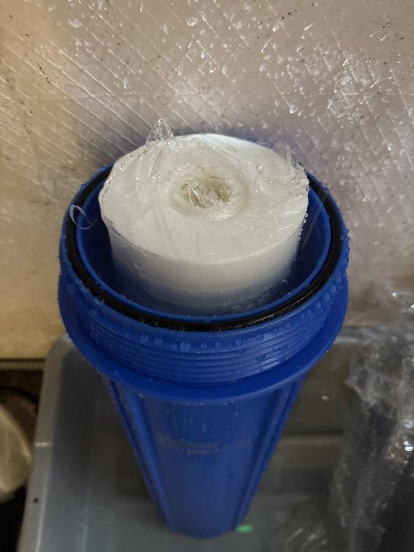 Inspected water filtration system and documented condition of blue filter housing with white pleated cartridge. Filter housing showed moisture accumulation and condensation on exterior surfaces. Client will contact to schedule warranty part installation once replacement component is received.