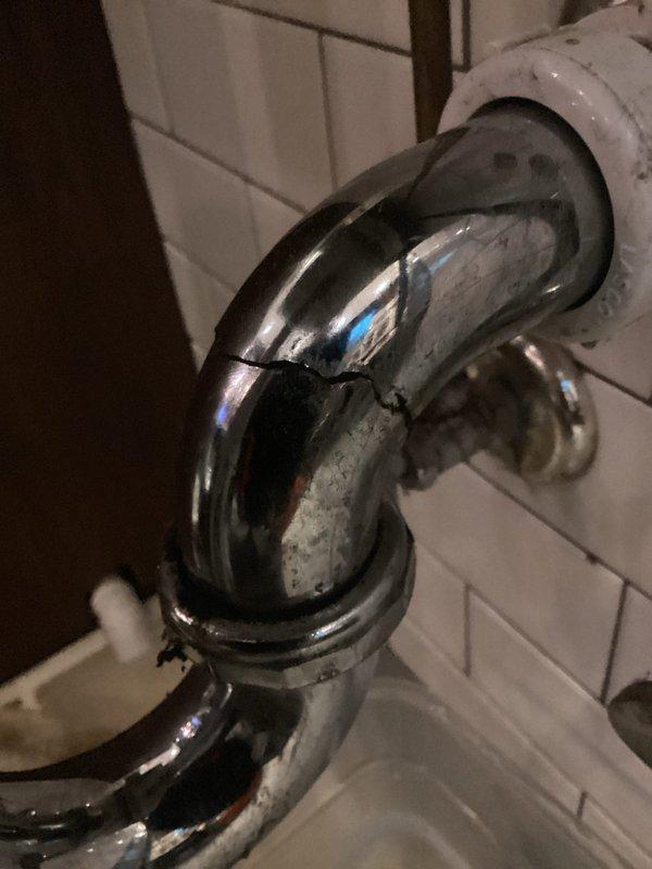 Responded to commercial plumbing service call for hand washing sink with cracked pipe. Upon inspection, identified deteriorated chrome P-trap assembly showing significant corrosion, water staining, and compromised structural integrity. Documented existing plumbing configuration including copper supply lines, chrome shutoff valves, and drain connections mounted against subway tile wall. Photographed damaged trap assembly and surrounding plumbing components to provide visual documentation of condition requiring repair or replacement.
