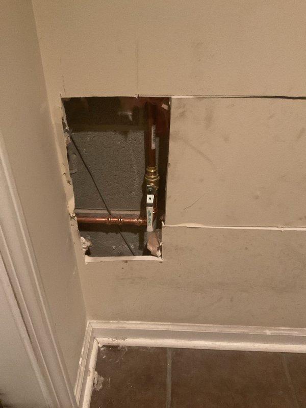 Installed new plumbing fixture with copper pipe connections behind drywall access point. Cut access panel in interior wall to route and connect copper supply lines using brass fittings and shutoff valve, with exterior green valve mounted on brick wall for main water control.