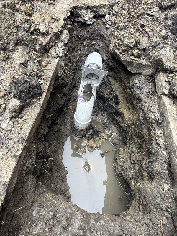 Completed sewer line installation with excavation and cleanout access point placement. Installed white PVC pipe fitting with removable cap to provide maintenance access to underground sewer system. Excavation revealed deteriorated concrete surrounding the work area, which was removed to facilitate proper pipe installation and ensure structural integrity of the new sewer line connection.