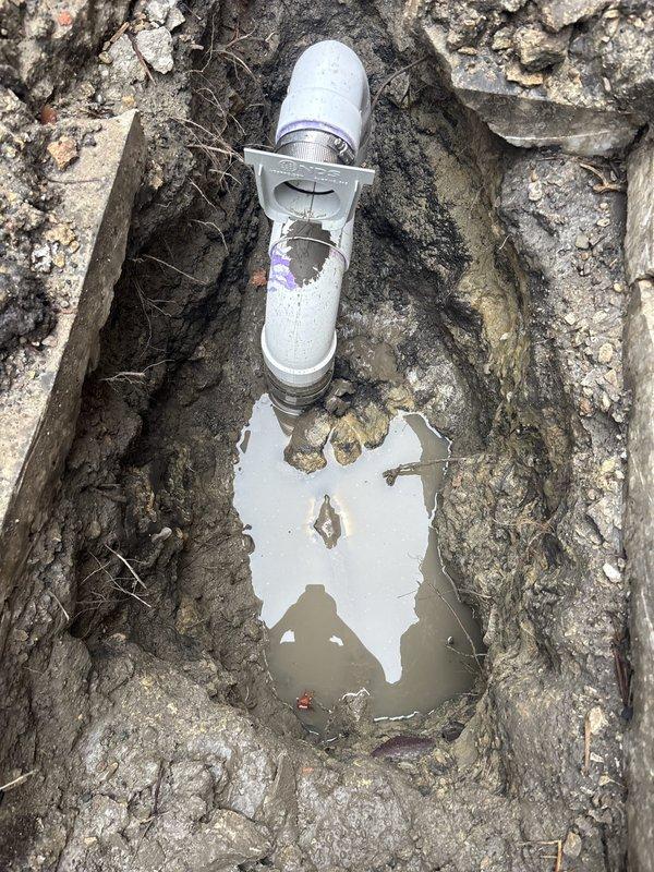 Completed sewer line installation with excavation and cleanout access point placement. Installed white PVC pipe fitting with removable cap to provide maintenance access to underground sewer system. Excavation revealed deteriorated concrete surrounding the work area, which was removed to facilitate proper pipe installation and ensure structural integrity of the new sewer line connection.