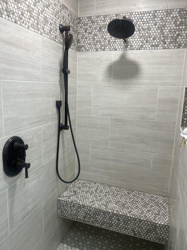 Responded to low water pressure complaint in modern walk-in shower equipped with dual shower system including ceiling-mounted rainfall showerhead and wall-mounted handheld unit with slide bar, all featuring matte black thermostatic valve controls. Upon inspection, identified issues with shower valve distribution and water supply lines affecting flow to both the overhead rainfall fixture and handheld shower components. Documented existing installation showing large-format tile work with decorative penny-round mosaic accents and professional plumbing fixture placement. Diagnosed distribution valve requiring repair to restore proper water pressure throughout the dual shower system. Captured detailed photographic documentation of shower configuration, fixture positioning, and tile work for reference. Scheduled follow-up appointment to provide repair estimates for shower valve and distribution water line corrections necessary to resolve pressure issues and restore full functionality to both