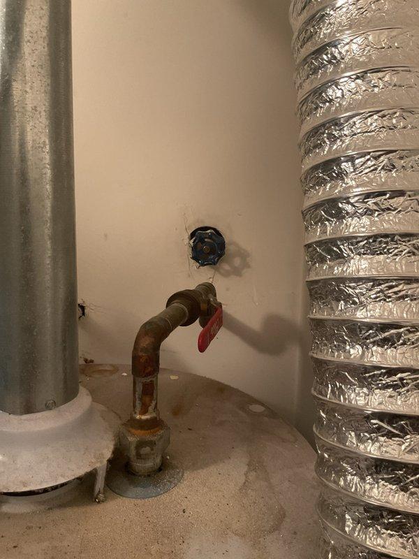 Conducted water heater assessment in utility closet and determined tank-style unit requires replacement due to age (10-15 years old) and visible corrosion on copper piping connections. Documented existing installation with corroded copper supply lines showing rust accumulation at base and oxidation throughout, red-wrapped insulation sections, and aging shut-off valve. Photographed utility space configuration including adjacent HVAC ductwork and laundry appliances to prepare comprehensive replacement estimate for new tank water heater installation.