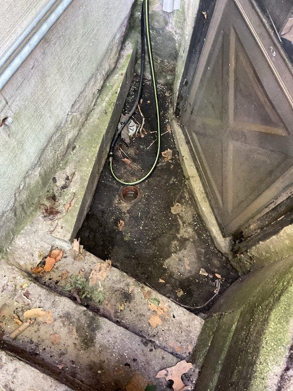 Responded to a clogged front deck drain at a century-old building. Upon arrival, found the exterior drainage channel heavily obstructed with organic debris including dead leaves, twigs, and decomposed plant matter, along with significant algae and moss growth on surrounding surfaces due to chronic moisture exposure. Cleared the drain opening and surrounding area of all debris, removed blockage from the drainage system, and verified proper water flow through the drain. The drainage channel showed evidence of long-term neglect with accumulated sediment and biological growth on concrete surfaces and exterior walls, which was contributing to the drainage issue.
