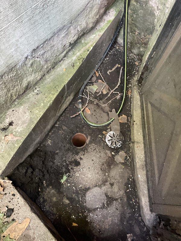 Responded to a clogged front deck drain at a century-old building. Upon arrival, found the exterior drainage channel heavily obstructed with organic debris including dead leaves, twigs, and decomposed plant matter, along with significant algae and moss growth on surrounding surfaces due to chronic moisture exposure. Cleared the drain opening and surrounding area of all debris, removed blockage from the drainage system, and verified proper water flow through the drain. The drainage channel showed evidence of long-term neglect with accumulated sediment and biological growth on concrete surfaces and exterior walls, which was contributing to the drainage issue.