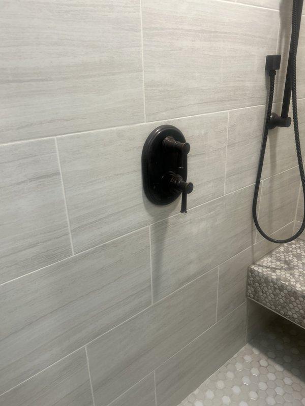 Completed installation of modern shower system featuring black matte finish fixtures including ceiling-mounted rainfall showerhead, handheld shower attachment with sliding bar, and dual-handle thermostatic valve control. Installed large-format rectangular tile in light gray with horizontal linear pattern on shower walls, with decorative hexagonal mosaic tile accent on built-in corner bench and shower floor for slip resistance and aesthetic appeal.