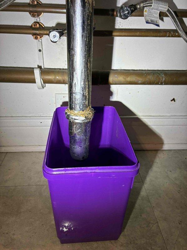 Responded to water leak in utility area where vertical galvanized steel drain pipe showed active leaking at connection point beneath sink basin. Upon inspection, identified significant corrosion and mineral deposit accumulation at pipe junction, with deteriorated galvanized finish indicating advanced age and wear. Temporary collection container was in place to catch dripping water. 

Documented extensive water staining and discoloration on underside of sink basin, confirming prolonged leak exposure. Examined surrounding copper supply lines and fittings, noting greenish patina consistent with aged plumbing system. Assessed drain assembly connections and identified compromised seal at pipe-to-basin junction as primary leak source. 

Provided photographic documentation of corroded pipe sections, mineral buildup, and water damage for estimate preparation. Recommended replacement of deteriorated galvanized drain pipe section and associated fittings to resolve active leak and prevent further