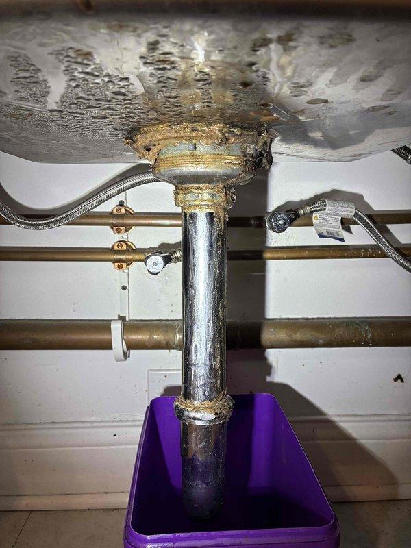 Responded to water leak in utility area where vertical galvanized steel drain pipe showed active leaking at connection point beneath sink basin. Upon inspection, identified significant corrosion and mineral deposit accumulation at pipe junction, with deteriorated galvanized finish indicating advanced age and wear. Temporary collection container was in place to catch dripping water. 

Documented extensive water staining and discoloration on underside of sink basin, confirming prolonged leak exposure. Examined surrounding copper supply lines and fittings, noting greenish patina consistent with aged plumbing system. Assessed drain assembly connections and identified compromised seal at pipe-to-basin junction as primary leak source. 

Provided photographic documentation of corroded pipe sections, mineral buildup, and water damage for estimate preparation. Recommended replacement of deteriorated galvanized drain pipe section and associated fittings to resolve active leak and prevent further