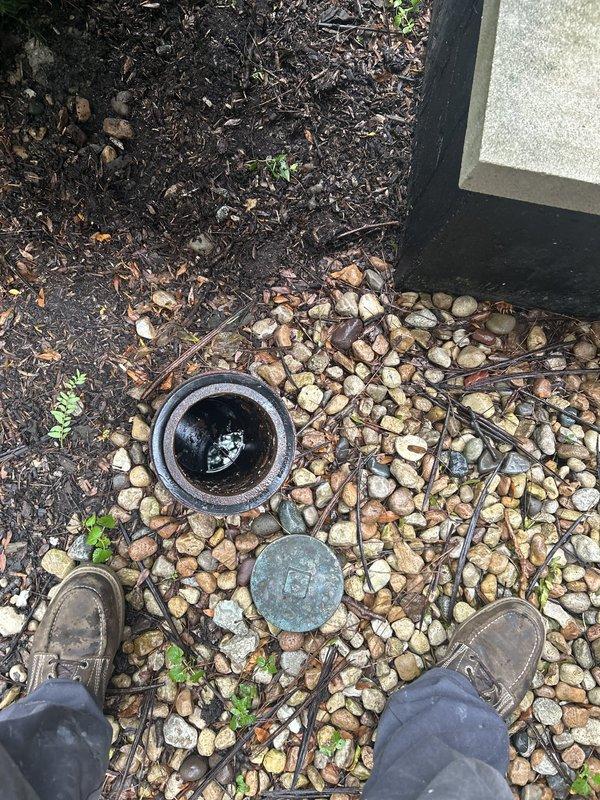 Responded to customer report of water intrusion during rainfall. Inspected exterior drainage system and located ground-level drain near building foundation with displaced access cover. Assessed door threshold area and surrounding hardscape for proper water management and drainage flow patterns to prevent water entry into unit.