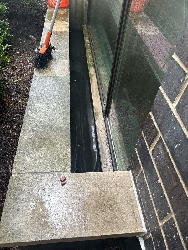 Responded to customer report of water intrusion during rainfall. Inspected exterior drainage system and located ground-level drain near building foundation with displaced access cover. Assessed door threshold area and surrounding hardscape for proper water management and drainage flow patterns to prevent water entry into unit.
