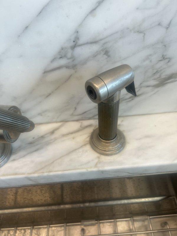 Installed Kohler Model 7038 water treatment system component with float valve assembly and connected to existing copper supply lines. Completed kitchen sink sprayer installation featuring brushed bronze finish sprayer head with ridged cylindrical base, coordinating with existing decorative faucet hardware on marble countertop.