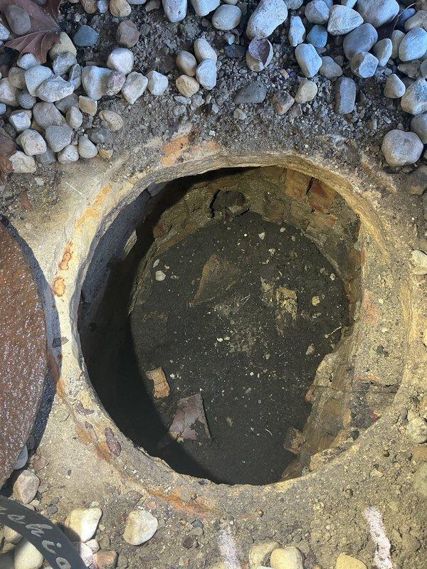 Responded to priority service call for failing catch basin in backyard of historic 130-year-old residence. Upon arrival, found circular catch basin with severely deteriorated concrete rim showing significant weathering, structural cracks, and brown/orange discoloration from prolonged moisture exposure and age. Basin contained standing water with sediment accumulation and debris, indicating compromised drainage function. The structure exhibited extensive surface deterioration around the circumference with the basin rim actively collapsing inward, creating a safety hazard in the landscaped area surrounded by river rock.

Assessed the structural integrity of the existing catch basin and determined complete replacement was necessary due to the advanced state of deterioration. Removed damaged concrete rim sections and deteriorated metal components visible in the work area. Excavated and cleared the basin opening, removing accumulated sediment, standing water, and organic debris from the vau