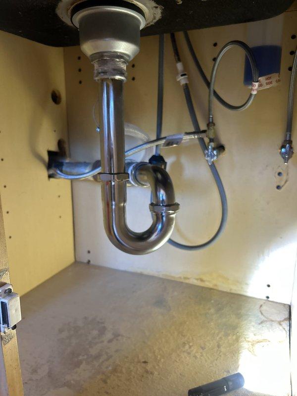 # Plumbing Service - Water Leak

Responded to a leaking kitchen sink in a 25-year-old residential property with original plumbing fixtures. Upon inspection, identified active water leak at the sink drain assembly beneath a stainless steel basin showing significant age-related wear and discoloration.

Accessed the cabinet interior and examined the chrome P-trap assembly and drain connections. Found water staining and moisture accumulation on the cabinet floor, indicating an ongoing leak from the drain system. The existing drain assembly consisted of a sink strainer, tailpiece extension, and U-shaped P-trap, all showing signs of deterioration consistent with the age of the plumbing system.

Inspected all visible supply lines, including flexible hoses and braided supply lines secured with metal fittings. Evaluated the integrity of all connections and seals throughout the drain assembly. Tested water flow to identify the exact source of the leak and assessed whether repair or replacement w