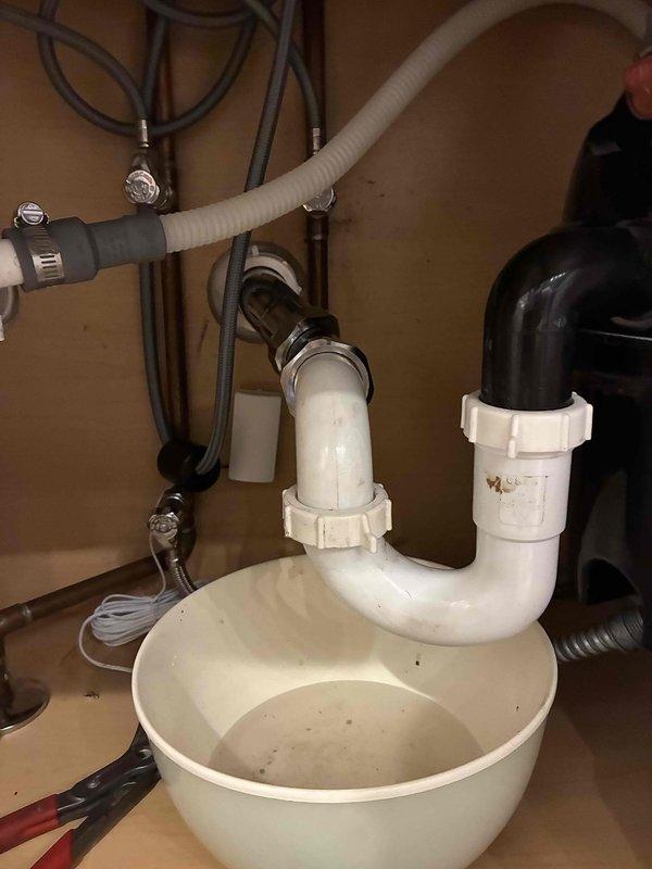 Responded to emergency call for water leak underneath kitchen sink in 19-year-old residential property. Upon arrival, inspected under-sink plumbing system and identified active leak at P-trap assembly. Found compromised compression fittings and deteriorated washers causing water to drip into cabinet space. 

Disassembled white PVC P-trap configuration and assessed all connection points. Replaced worn compression fittings and rubber washers at trap connections. Verified proper alignment of drain piping from sink basin to main drain line. Inspected adjacent garbage disposal unit connections and black corrugated drain hose for any secondary leak sources. Reassembled P-trap assembly with new components, ensuring proper seal at all joints. Tested system by running water through sink and disposal unit, confirming no further leakage. Cleaned cabinet interior and removed temporary collection basin. All plumbing components functioning properly upon completion.