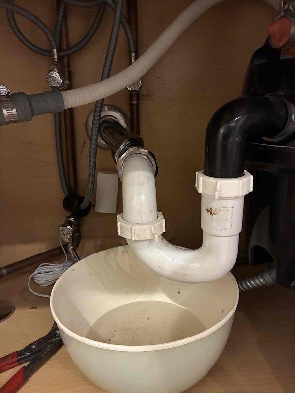 Responded to emergency call for water leak underneath kitchen sink in 19-year-old residential property. Upon arrival, inspected under-sink plumbing system and identified active leak at P-trap assembly. Found compromised compression fittings and deteriorated washers causing water to drip into cabinet space. 

Disassembled white PVC P-trap configuration and assessed all connection points. Replaced worn compression fittings and rubber washers at trap connections. Verified proper alignment of drain piping from sink basin to main drain line. Inspected adjacent garbage disposal unit connections and black corrugated drain hose for any secondary leak sources. Reassembled P-trap assembly with new components, ensuring proper seal at all joints. Tested system by running water through sink and disposal unit, confirming no further leakage. Cleaned cabinet interior and removed temporary collection basin. All plumbing components functioning properly upon completion.