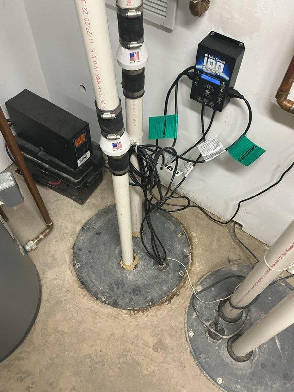 Installed comprehensive dual sump pump system with electronic monitoring and control equipment in basement utility room. System includes two white PVC discharge pipes with rubber couplings emerging from concrete floor pits with metal covers, two wall-mounted black control boxes with digital displays for pump operation and backup monitoring, and integrated wiring for power and communication between components. All components properly secured to walls and floor, with copper supply piping connected to existing plumbing infrastructure and expansion tank mounted for system pressure regulation.