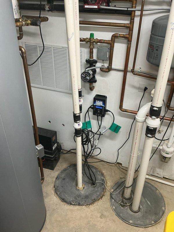 Installed comprehensive dual sump pump system with electronic monitoring and control equipment in basement utility room. System includes two white PVC discharge pipes with rubber couplings emerging from concrete floor pits with metal covers, two wall-mounted black control boxes with digital displays for pump operation and backup monitoring, and integrated wiring for power and communication between components. All components properly secured to walls and floor, with copper supply piping connected to existing plumbing infrastructure and expansion tank mounted for system pressure regulation.