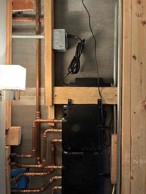Completed installation of radiant floor heating manifold system with copper piping configuration and electronic control panel in mechanical room. Installed modern glass-enclosed shower fixture with black metal frame, marble-look tile surround, and mosaic floor tile with integrated drain system. System requires scheduling for final connection and testing before activation.