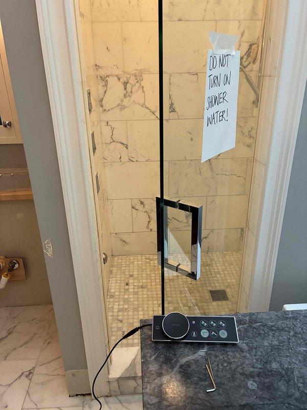 Completed installation of radiant floor heating manifold system with copper piping configuration and electronic control panel in mechanical room. Installed modern glass-enclosed shower fixture with black metal frame, marble-look tile surround, and mosaic floor tile with integrated drain system. System requires scheduling for final connection and testing before activation.