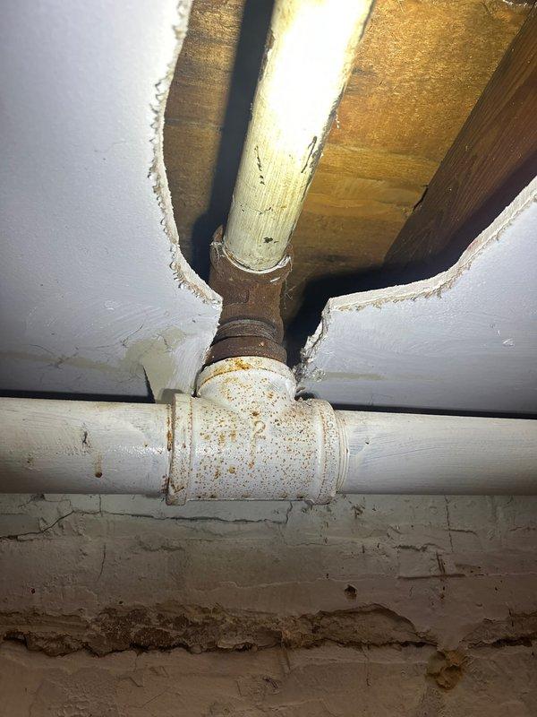 Completed installation of PVC piping junction in basement crawl space, including vertical riser and horizontal run with T-fitting connections. Addressed existing corrosion and rust staining around pipe fittings and joints, ensuring proper alignment through floor joists and structural openings. Documentation photos captured for quality assurance and future reference.