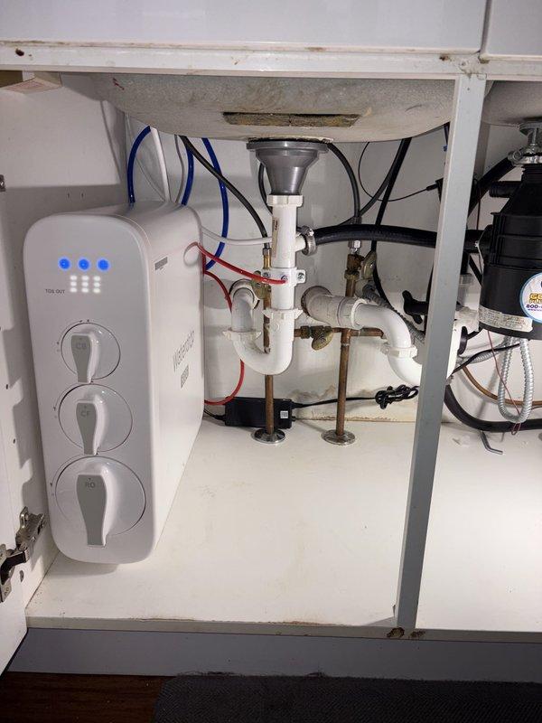 Installed tankless water filtration system under kitchen sink with undermount stainless steel basin and brushed metal single-handle faucet. Mounted TCM-WH electronic control unit to interior cabinet wall with three LED indicators and control buttons, integrated system with existing white PVC P-trap drain configuration and garbage disposal unit. Connected blue supply lines, electrical wiring, and plumbing connections to complete installation on granite countertop with matching backsplash and white gloss cabinetry.