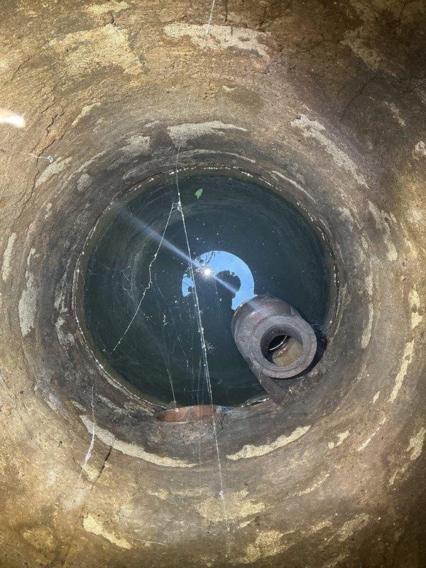 Completed residential drain and sewer estimate involving inspection of well/cistern system and attic plumbing infrastructure. Documented deep cylindrical concrete well showing significant weathering, moisture staining, and mineral deposits along interior walls, with standing water at bottom and visible debris including submerged rubber component. Photographed attic space containing exposed plumbing lines, electrical conduits, and aged structural elements requiring assessment for potential sewer line routing or repair access.