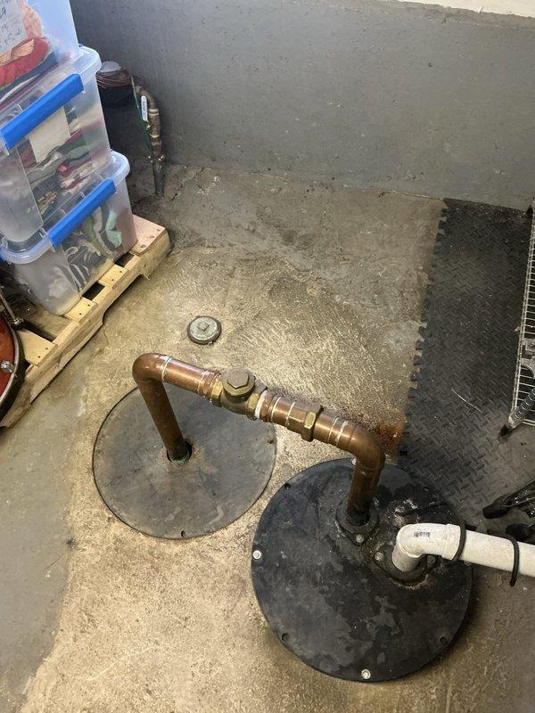 Conducted residential drain and sewer system estimate in utility room with focus on existing sump basin and drainage infrastructure. Documented aging copper and brass piping configuration emerging from floor access points, along with PVC drain connections showing signs of oxidation and mineral buildup. Evaluated sump pit condition, noting stagnant water with visible debris, corroded pipe fittings, and sediment accumulation indicating potential drainage concerns requiring system assessment and recommendations for repair or replacement.