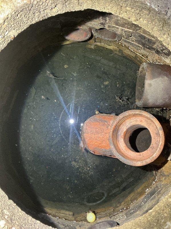 Conducted residential drain and sewer system estimate in utility room with focus on existing sump basin and drainage infrastructure. Documented aging copper and brass piping configuration emerging from floor access points, along with PVC drain connections showing signs of oxidation and mineral buildup. Evaluated sump pit condition, noting stagnant water with visible debris, corroded pipe fittings, and sediment accumulation indicating potential drainage concerns requiring system assessment and recommendations for repair or replacement.