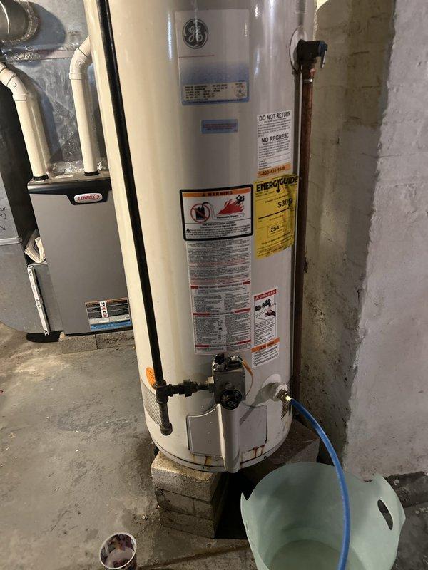 Responded to service call for water heater leaking at base. Upon inspection, found GE brand tanked water heater installed in 2010 showing active leak at bottom of unit. Documented existing plumbing configuration including red-handled shut-off valve, metal supply lines, and flexible connectors mounted to floor joists. Photographed current installation setup and leak condition. Provided estimate for water heater replacement due to tank failure, as leaks at base typically indicate internal tank corrosion that cannot be repaired.
