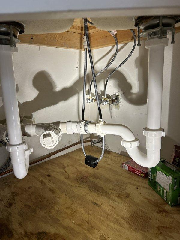 Responded to service call for kitchen plumbing issues involving a malfunctioning faucet and stuck water shutoff valve. Upon inspection, found existing kitchen faucet was improperly installed backwards, exhibiting active leaks and general disrepair requiring full replacement. Assessed under-sink plumbing configuration featuring double-basin setup with white PVC P-trap assemblies and braided stainless steel supply lines connected to chrome shutoff valves mounted on cabinet back wall. Evaluated stuck water valve that would not close properly, determining it required careful assessment to avoid forcing and potentially damaging the valve mechanism. Provided estimate for replacement of kitchen faucet with customer-supplied fixture and repair or replacement of compromised shutoff valve as needed to restore proper water control functionality.