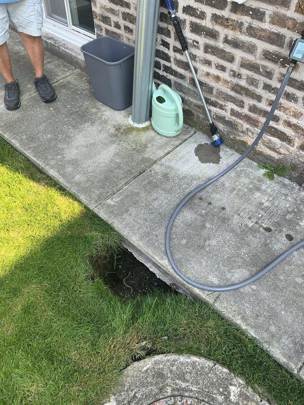 Responded to a plumbing service call at a residential property to address an outdoor drainage issue. Upon arrival, identified a ground-level access point or drainage opening located at the junction between the concrete patio and lawn area. Inspected the cavity and surrounding area, noting exposed soil and potential water infiltration concerns. Documented the existing conditions of the outdoor plumbing infrastructure, including hose connections and drainage pathways along the patio perimeter. Completed photographic documentation of the work area for service records.