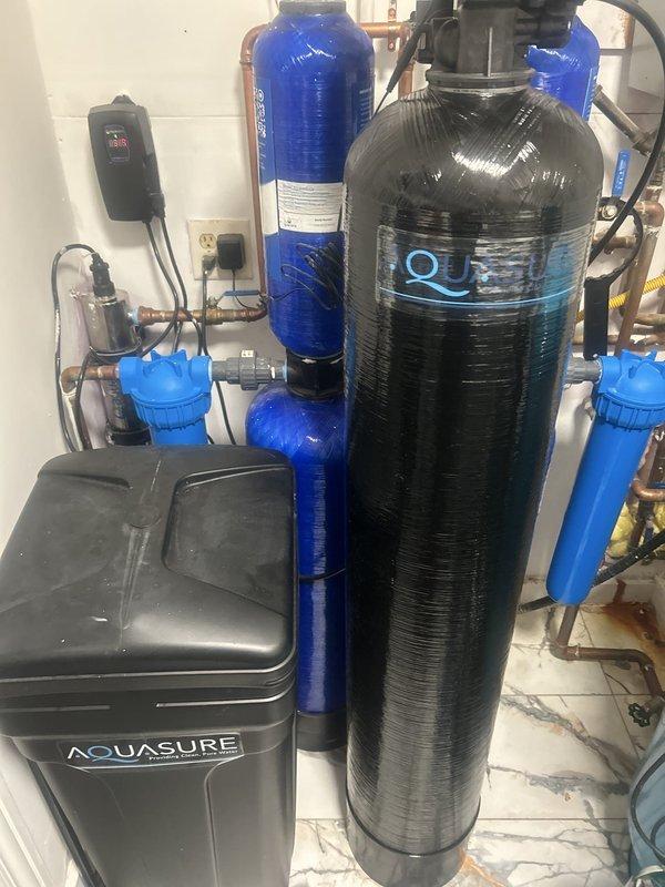 Responded to service call for malfunctioning water filtration and softening system at non-owner occupied property. Upon arrival, inspected comprehensive water treatment setup including Aquasure water softener with brine tank, multiple cylindrical filter housings with blue cartridge holders, and associated plumbing connections. Evaluated tankless water heater integration with treatment system, examining copper and PVC piping configurations, valve operations, and electrical control panel functionality. Diagnosed system performance issues, verified proper flow through filter housings and softener tank, checked brine tank levels and settings on digital control display. Restored water treatment system to proper working condition and documented installation with photographs for service records.